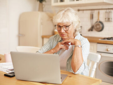 Modern technology, old people and retirement. Gray haired female pensioner in glasses filling in loan application form online, sitting in front of laptop, reading information with serious focused look